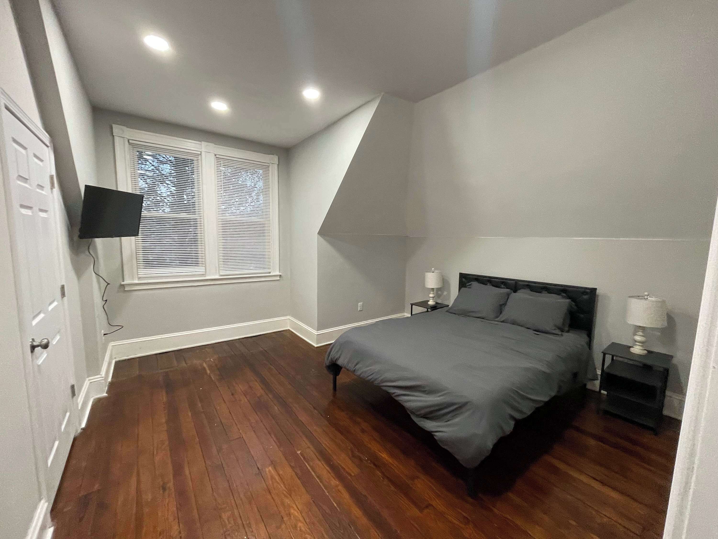 A bedroom with a bed, a TV mounted on the wall, a window with blinds, and a wooden floor.