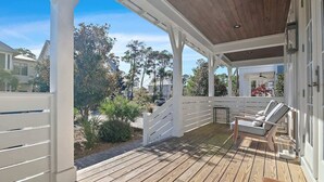 A covered outdoor deck with a white railing, a wooden floor, and a lounge chair. - Community pool, 4 Bikes 'Seas the Day' (Santa Rosa Beach)