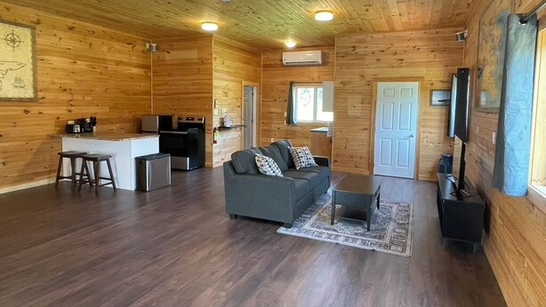 A cozy cabin interior with a wooden interior, a sofa, a coffee table, a kitchen area, and a television. - Tidal's Edge Cottage (Fortune Bridge)