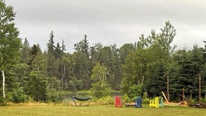 A grassy field with a hammock, colorful Adirondack chairs, and a forest in the background. - Tidal's Edge Cottage (Fortune Bridge)