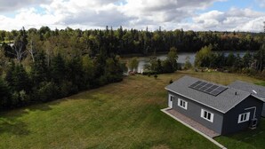 A house with a solar panel roof, surrounded by a large grassy yard and a forested area with a lake in the background. - Tidal's Edge Cottage (Fortune Bridge)