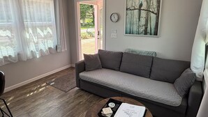 A living room with a grey sofa, a coffee table with a book and remote, a clock, and a painting of a forest.