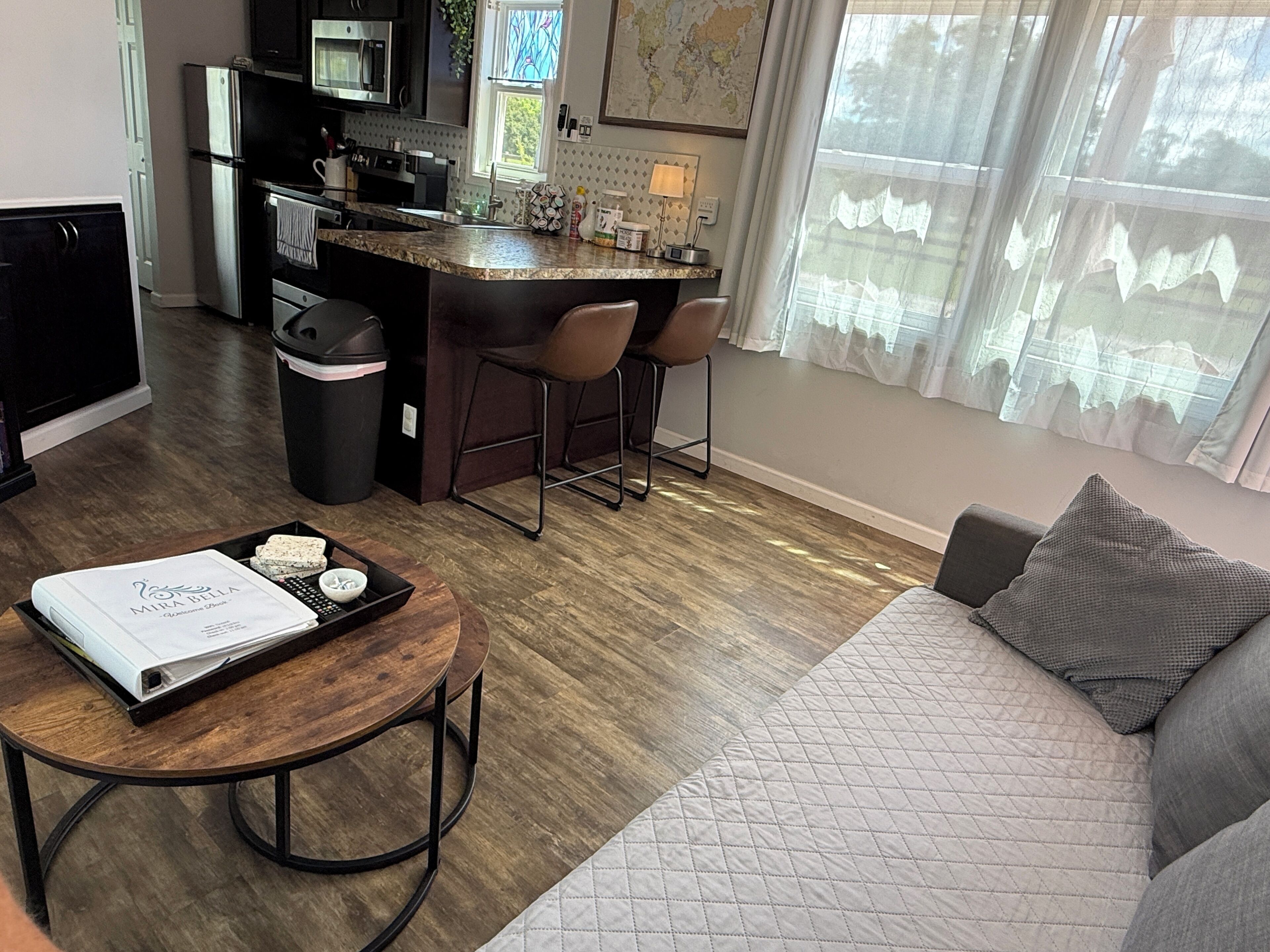 A modern kitchen with a central island, bar stools, a dining table with a tray, a sofa, and a window with curtains.