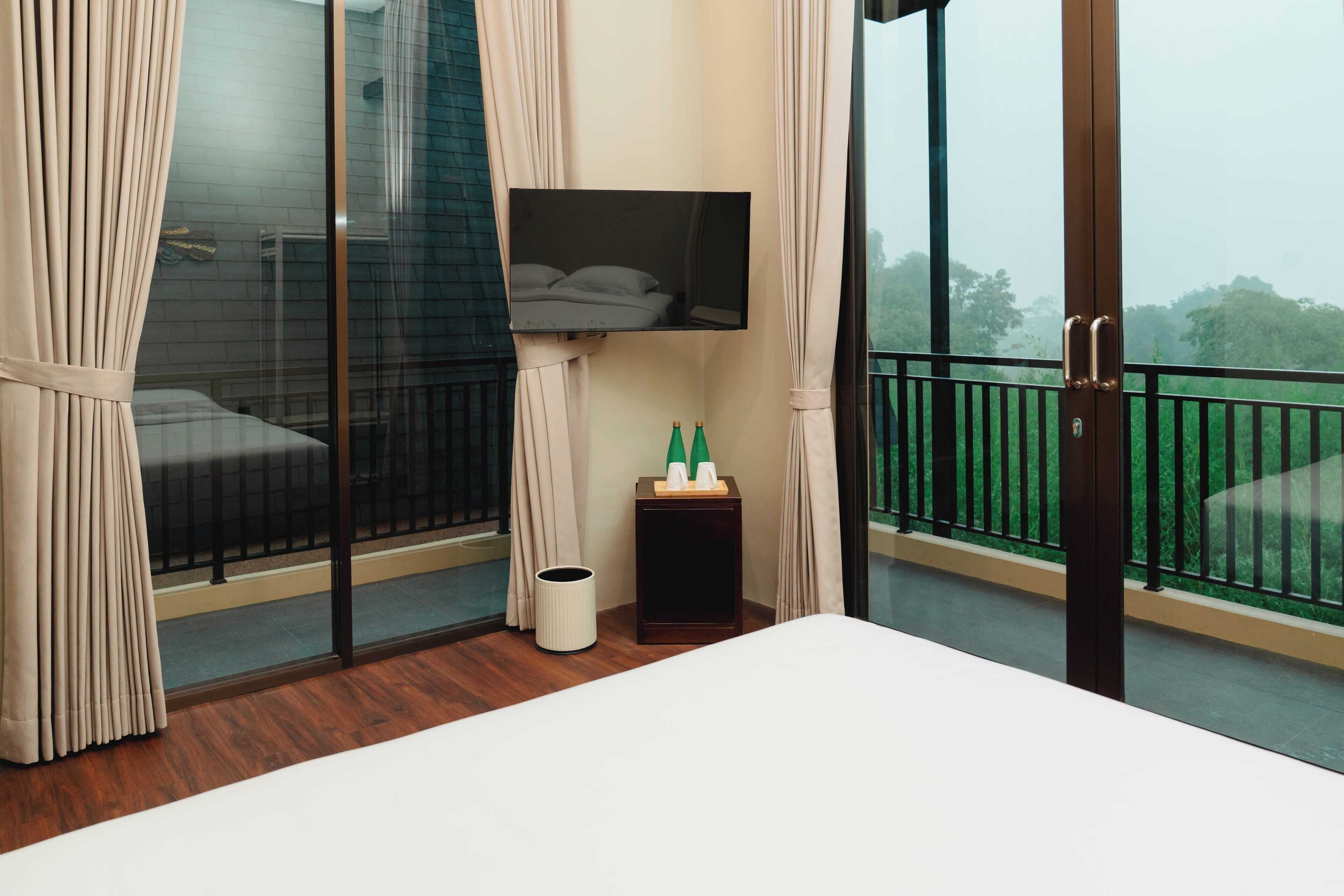 Habitación de hotel con una cama, una mesita con artículos de aseo, un televisor y un balcón con vistas a zonas verdes.