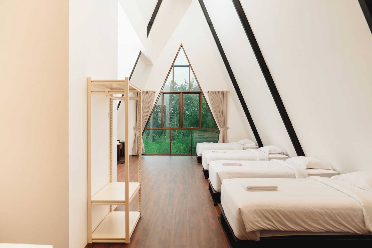 A modern, triangular-roofed room with wooden flooring, white beds, and a large window offering a view of greenery.