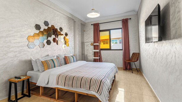 A bedroom with a bed, a wooden nightstand, a wall-mounted TV, and a window with curtains. - Massira Social Living (Saâda)