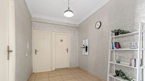 A hallway with a white door, a white shelf with books and a plant, and a clock on the wall. - Massira Social Living (Saâda)