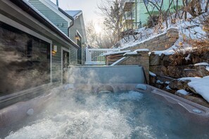 Een hottub met een stenen muur en een houten terras, omgeven door met sneeuw bedekte grond en bomen.