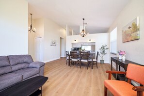 A spacious living room with a grey sofa, a wooden dining table with chairs, a kitchen in the background, and a chandelier.