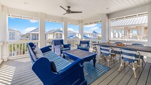 A covered outdoor seating area with blue furniture, a ceiling fan, and a view of houses in the background. - 🌅 Outdoor Oasis - Grill & TV | Private Beach | Bikes | Resort Pool ⛱️ (Seacrest beach)