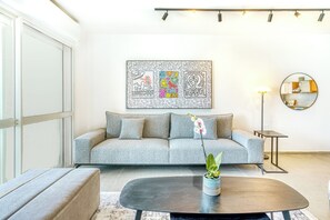 A modern living room with a grey sofa, a round coffee table with a plant, a wall-mounted artwork, a floor lamp, and a round mirror reflecting a kitchen.