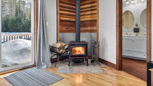 A room with a wood-burning stove, a large window with a view of a snowy landscape, and a wooden wall panel. - Hot Tub Lakeside Cottage – 30 Min from Mont Tremblant (Labelle)