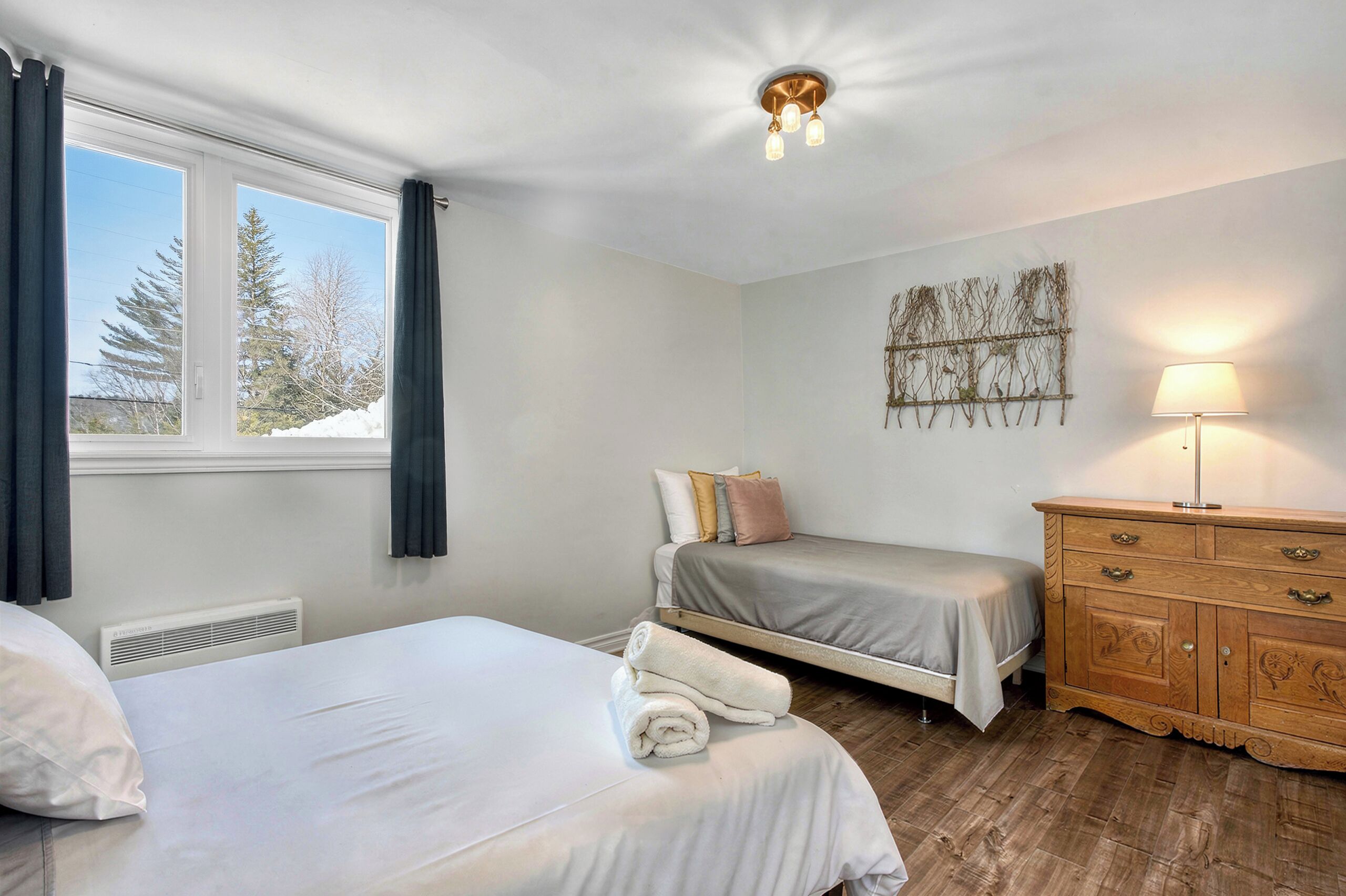 Une chambre avec deux lits, une commode, une lampe fixée au mur et une fenêtre avec des rideaux.