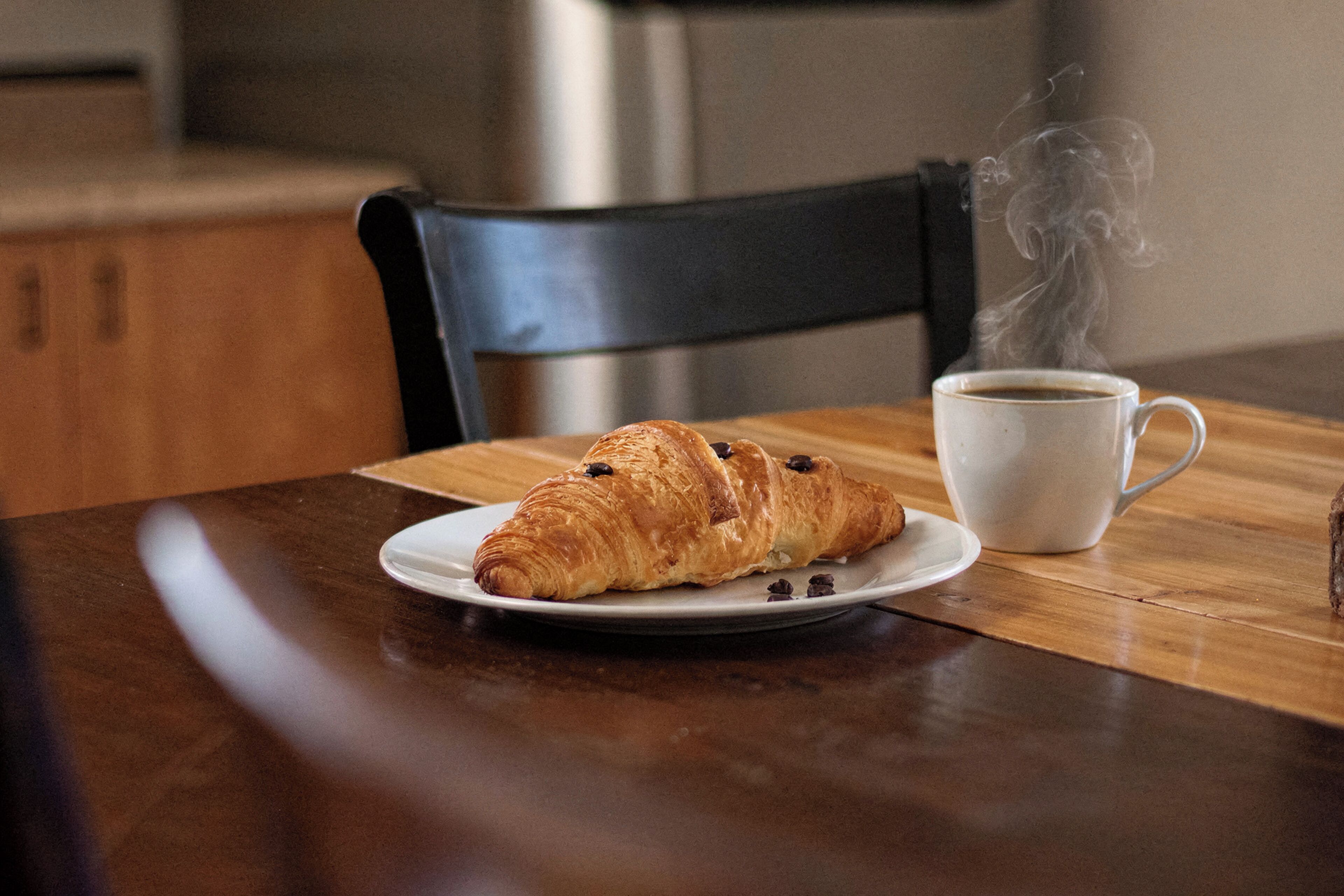 Um croissant num prato, uma chávena de café e uma cadeira sobre uma mesa de madeira.