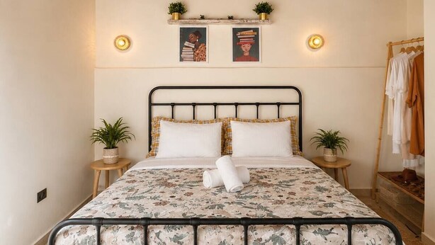 Una cama con colcha estampada, dos mesitas de noche con plantas, una repisa de pared con objetos decorativos y una lámpara colgante.