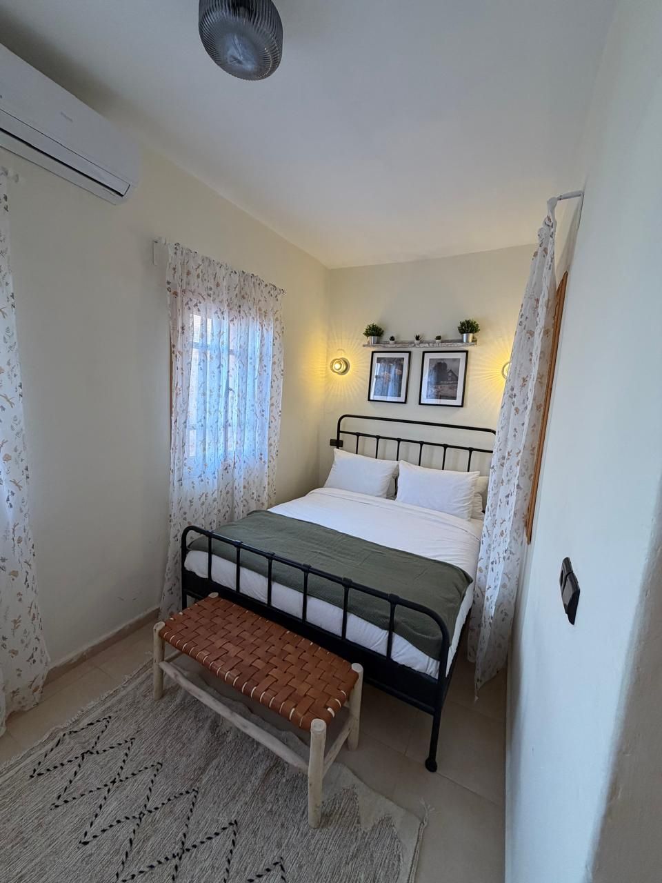 Quarto com cama, um banco, prateleira na parede com plantas e janela com cortinas.