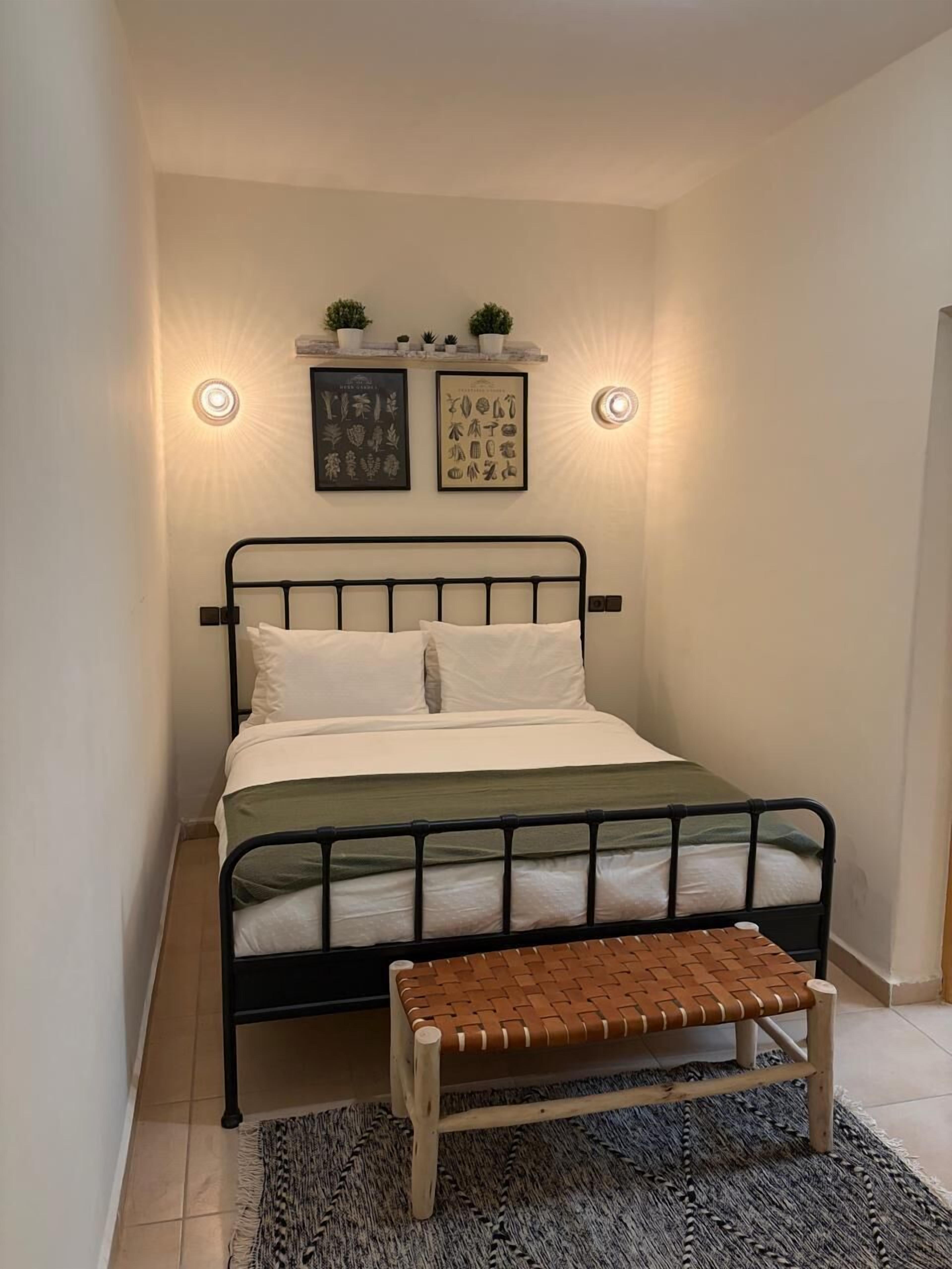 Quarto pequeno e bem arrumado, com uma cama, um banco e duas luminárias fixadas na parede.