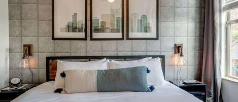 A hotel room with a bed, framed cityscape pictures, two bedside lamps, and a window with curtains.