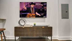 Ein Zimmer mit an der Wand befestigtem Fernseher, einer Holzkommode mit Pflanze darauf und einer Steckdose an der Wand.