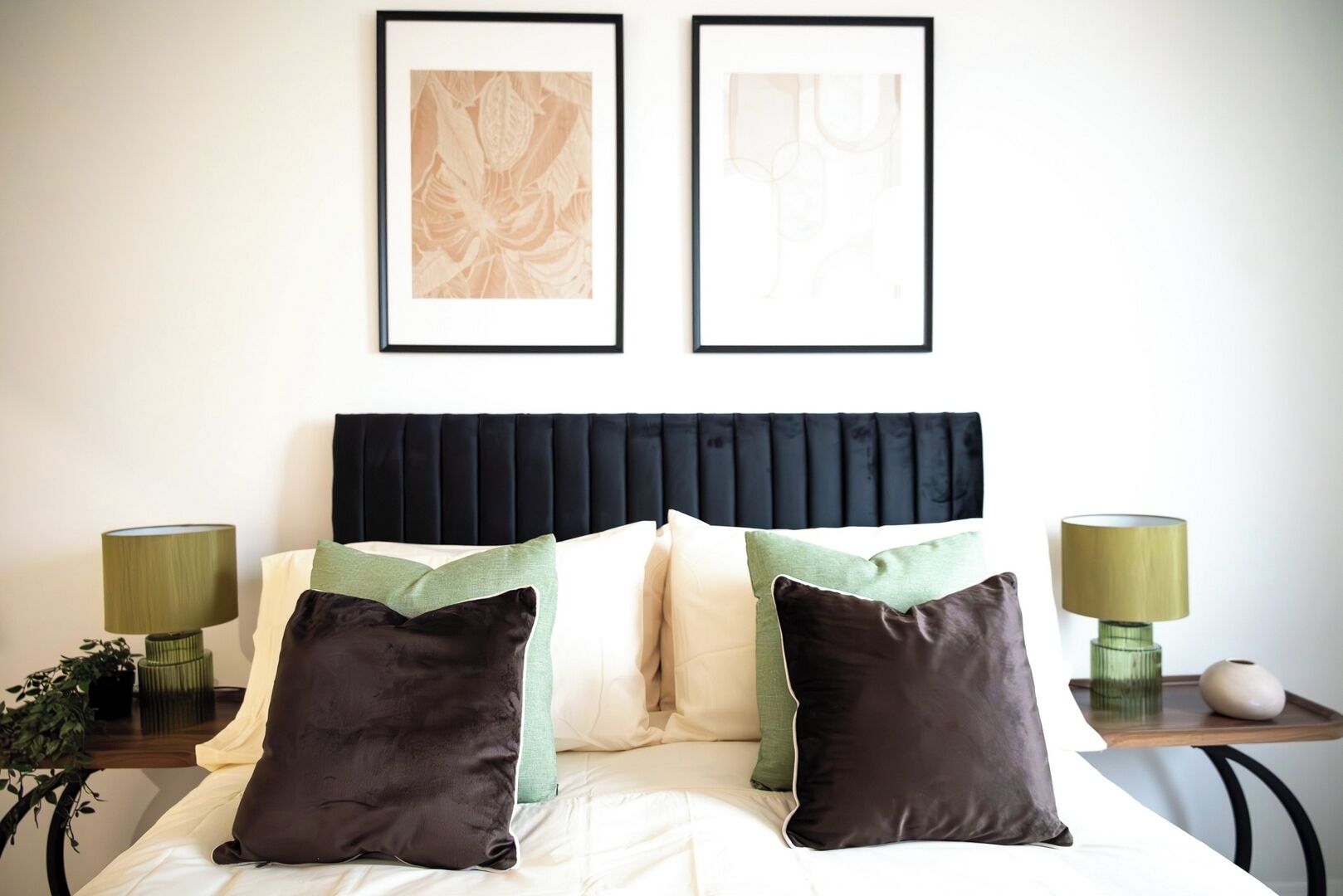 A neatly made bed with a black headboard, two framed abstract artworks above it, and two bedside tables with lamps and decorative items.