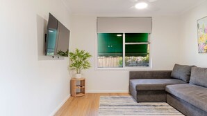 A modern living room with a sectional sofa, a flat-screen TV mounted on the wall, a potted plant, and a window with a view of greenery. - Botanic Base • Pets Welcome • Wagga Wagga (Mount Austin)