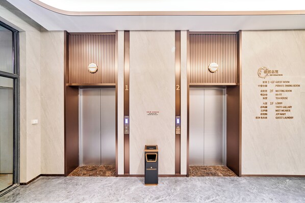 Un ascensor con dos puertas, cada una marcada con un número. Hay una pantalla digital encima de cada puerta, y un panel pequeño debajo de ellas.