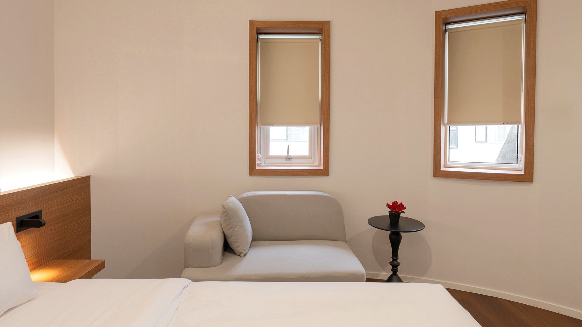 A hotel room with a bed, a sofa, a small table with a red flower, and two windows with blinds.