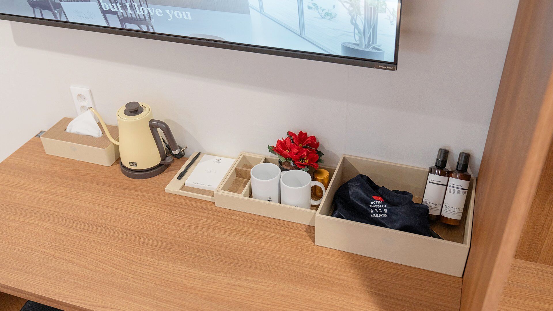 A hotel room with a wooden desk, a kettle, a tissue box, a tray with mugs, a potted plant, a bag, and bottles of lotion.