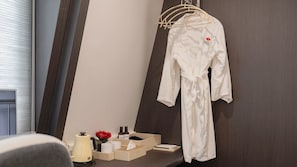 Habitación de hotel con una bata colgada en un perchero de madera, una mesita de noche con un hervidor de agua, un florero con flores rojas y una caja de artículos de aseo.