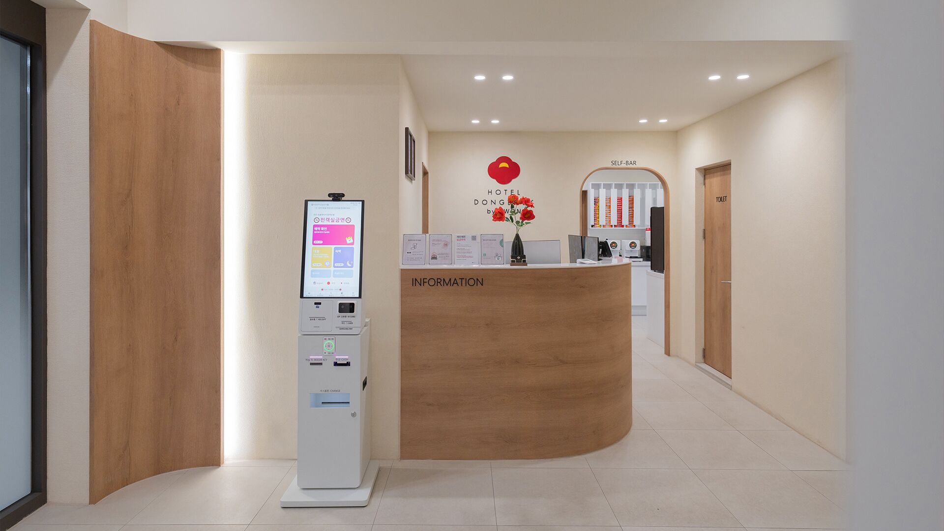 Et hotelreceptionsområde med en selvbetjeningskiosk, en træreception og et skilt, der angiver en selvbetjeningsbar.