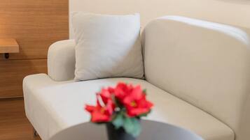 Un sofá moderno blanco con un solo cojín blanco, una mesita redonda con un florero de flores rojas y una mesita de madera.