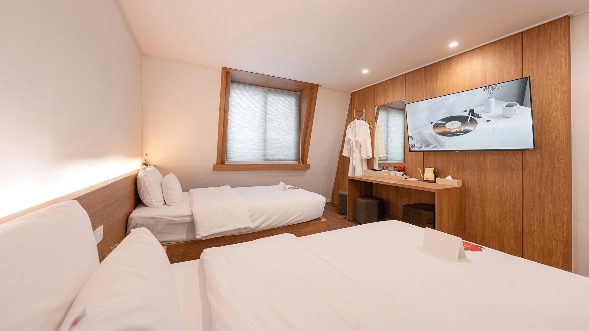 A hotel room with two beds, a wooden wardrobe, a desk with a mirror, and a large window with blinds.