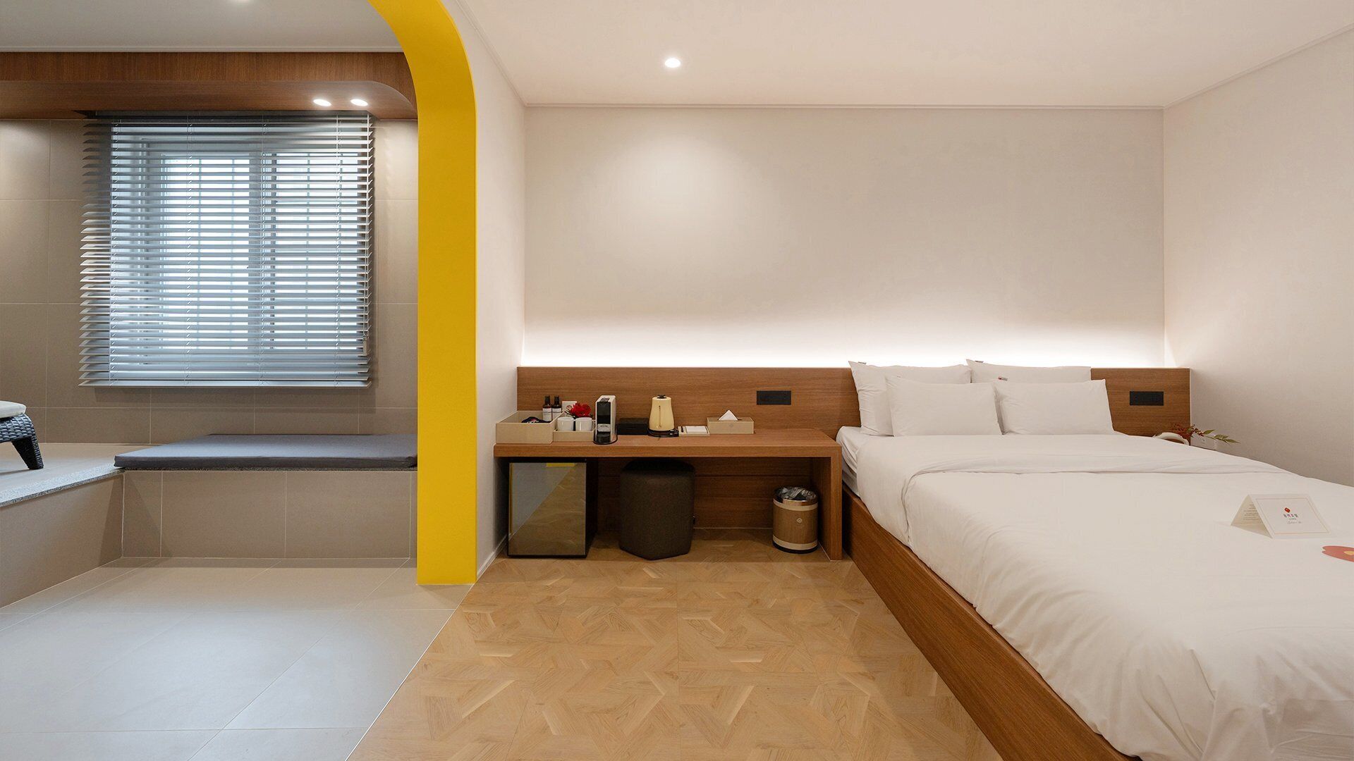 Habitación de hotel con cama, mesita de noche, ventana con persianas y un pequeño cubo de basura.