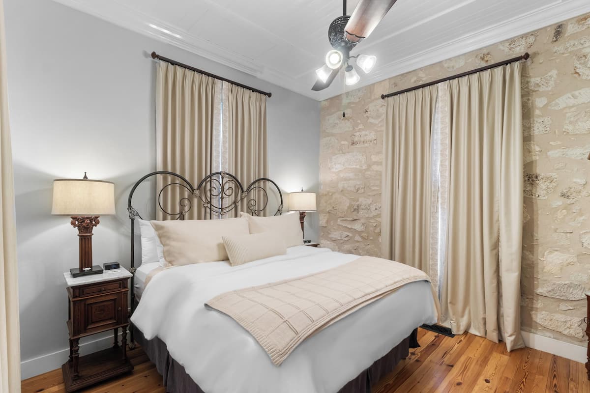 A bedroom with a bed, curtains, a nightstand with a lamp, and a stone wall.