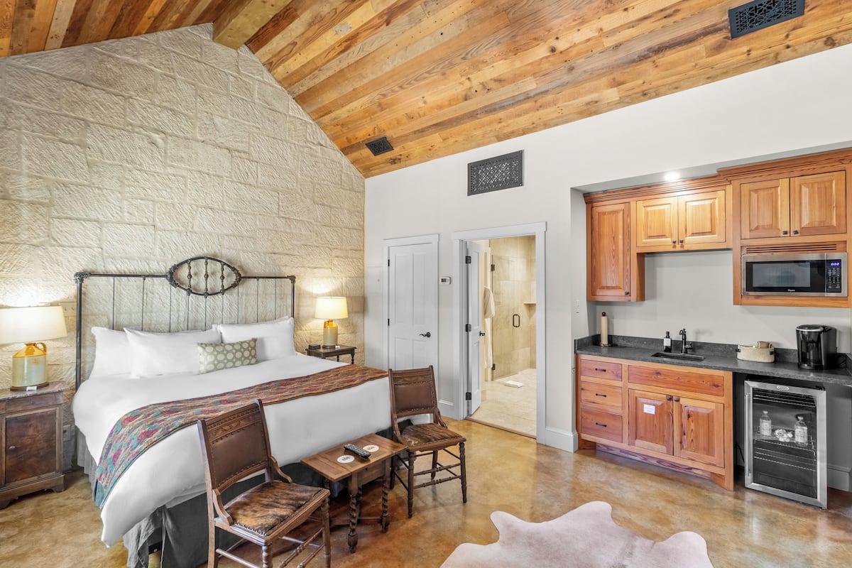 A bedroom with a large bed, wooden headboard, bedside tables, and a small table with chairs. Adjacent is a kitchen with wooden cabinets, a microwave, and a refrigerator.