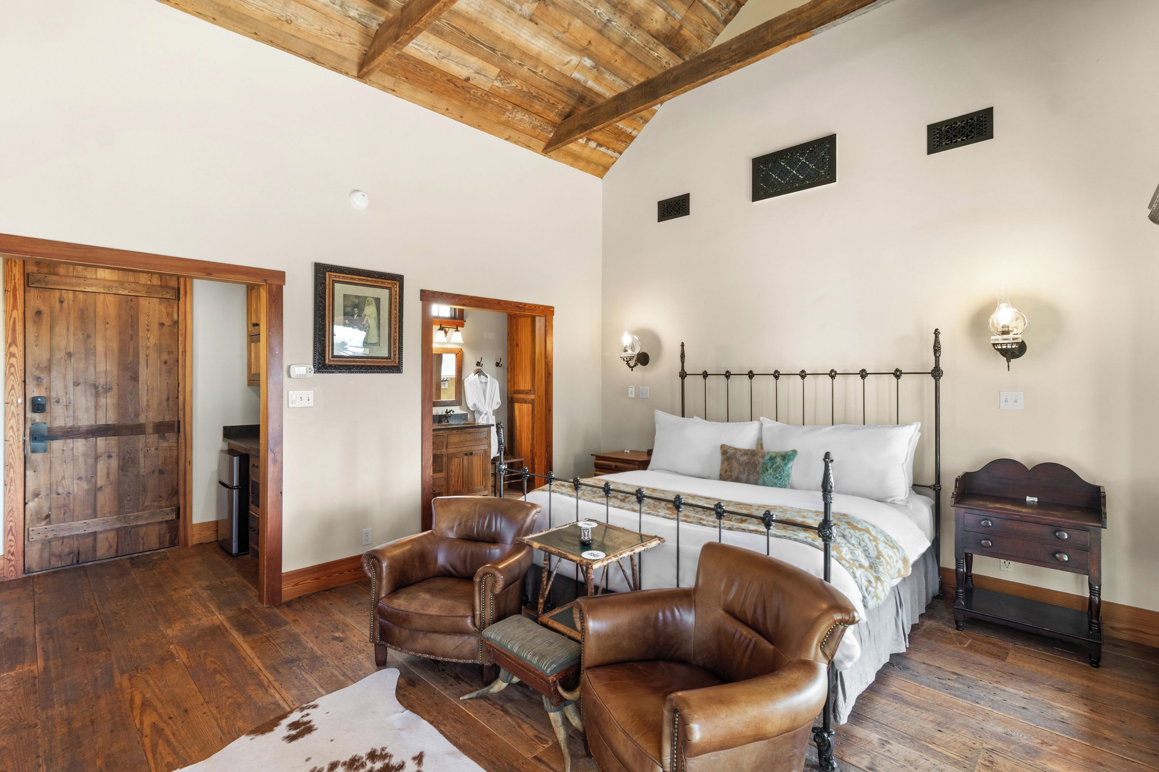 A bedroom with a wooden ceiling, a bed with a metal frame, two leather chairs, a small table, and a nightstand.