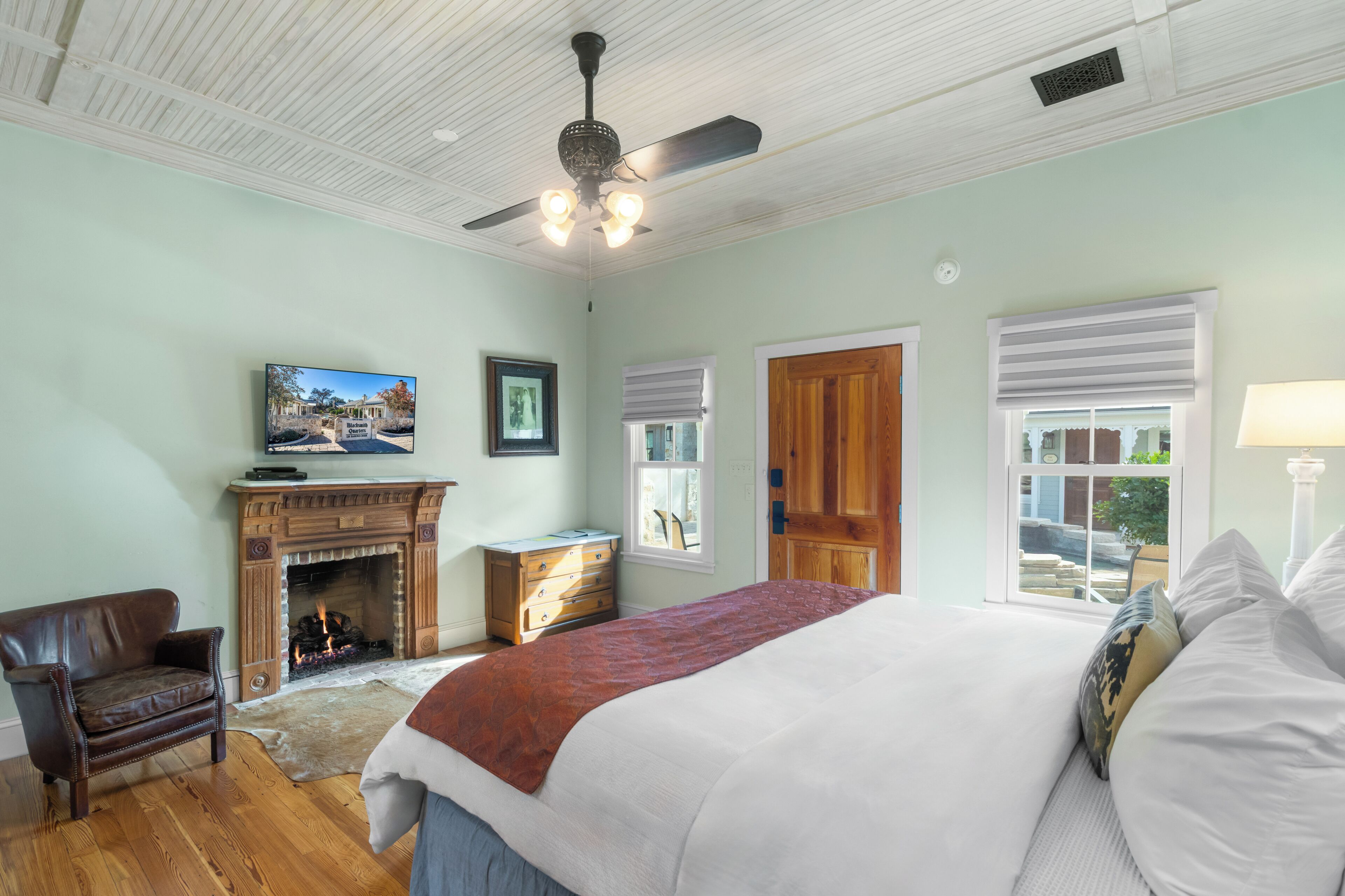 Une chambre avec une cheminée, un lit recouvert d’une couverture rouge, une porte en bois et un ventilateur de plafond.