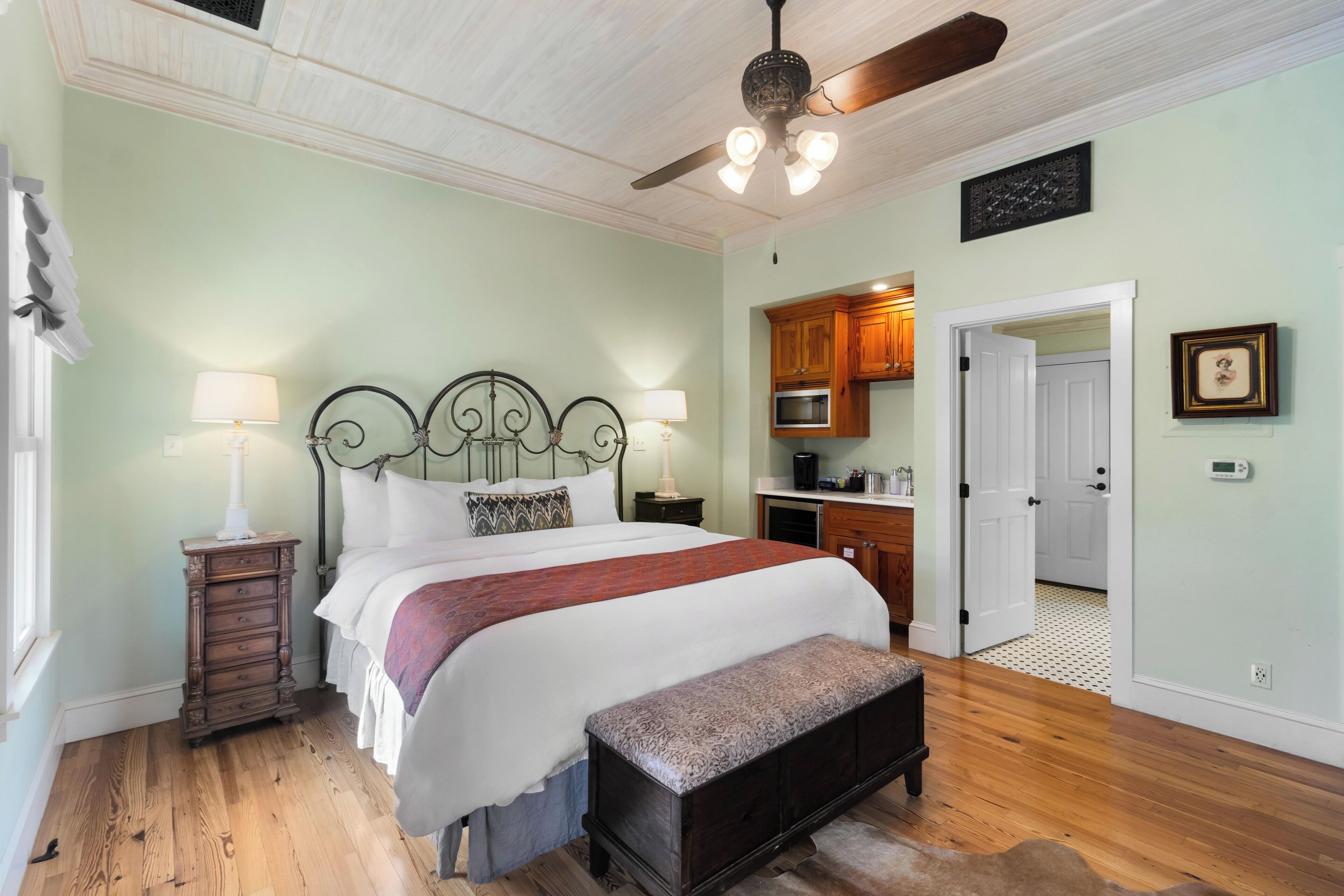 A bedroom with a bed, bedside tables, a bench, a ceiling fan, and a view into the kitchen.