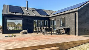 Ein modernes Haus mit einer Terrasse, einem großen Fenster und Solaranlagen auf dem Dach.