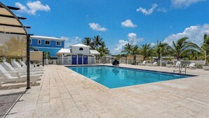 Una piscina con bordo piastrellato, un piccolo edificio con porte blu e palme sullo sfondo.