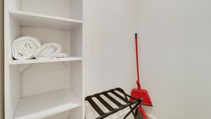 Un espacio de almacenamiento blanco con estantes, uno de los cuales tiene toallas enrolladas. Una escoba roja está apoyada contra la pared.