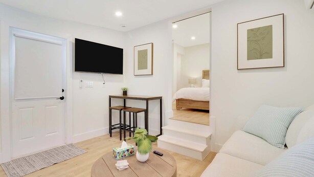 Una habitación moderna y bien iluminada con un televisor de pantalla plana, una mesa redonda con una planta, una pequeña mesa con dos sillas, una cama con una mesita de noche y obras de arte enmarcadas en las paredes.