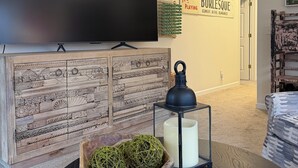 Una sala de estar moderna con un televisor de pantalla plana, una pared decorativa con un letrero, un mueble de madera, una mesa redonda con un farol y una zona de estar acogedora.