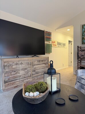 A modern living room with a flat-screen TV, a decorative wall with a sign, a wooden cabinet, a round table with a lantern, and a cozy seating area. - Acorn Cottage a Peaceful 3-bedroom house 1 mile from Elon University (Elon)