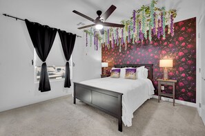 Een slaapkamer met een bed, een plafondventilator, een raam met gordijnen, een nachtkastje met een lamp en bloemenbehang.