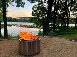 A metal fire pit with flames, surrounded by gravel, with a lake and trees in the background.