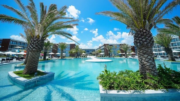 Ein Poolbereich mit Palmen, blauem Himmel und modernen Gebäuden im Hintergrund.