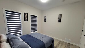 Ein Schlafzimmer mit einem Bett, zwei Fenstern mit Jalousien, einer Deckenleuchte und einem Lüftungsgitter.