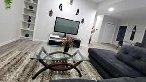 Ein modernes Wohnzimmer mit einem Glastisch, einem Flachbildfernseher, einem grauen Sofa und Dekoration auf Regalen.