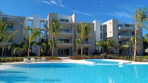 Een modern hotel met een zwembad, palmbomen en een helderblauwe lucht.